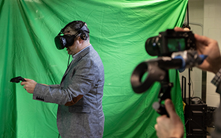 Dr. Adam Sachs stands in front of a green background wearing virtual reality head gear and holding virtual reality wands.